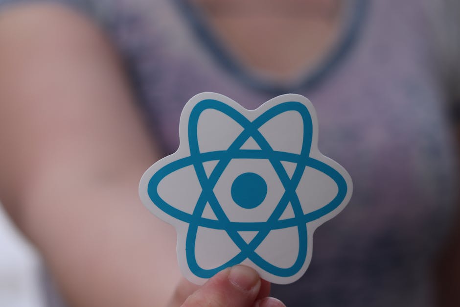 A person holding a small sticker featuring a stylized blue atomic symbol with intersecting orbits and a central dot, in front of a blurred background that includes a person wearing a purple top. The s
