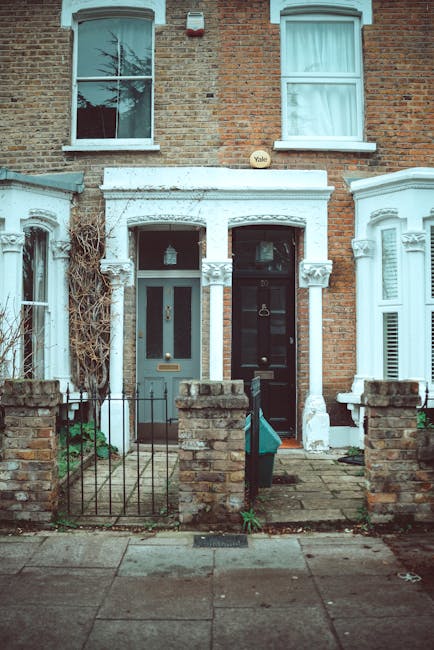 A row of residential terraced houses on a quiet Belsize Village street with cobblestone pavement, featuring brick and stucco facades painted in white and various shades of brown. The houses have small